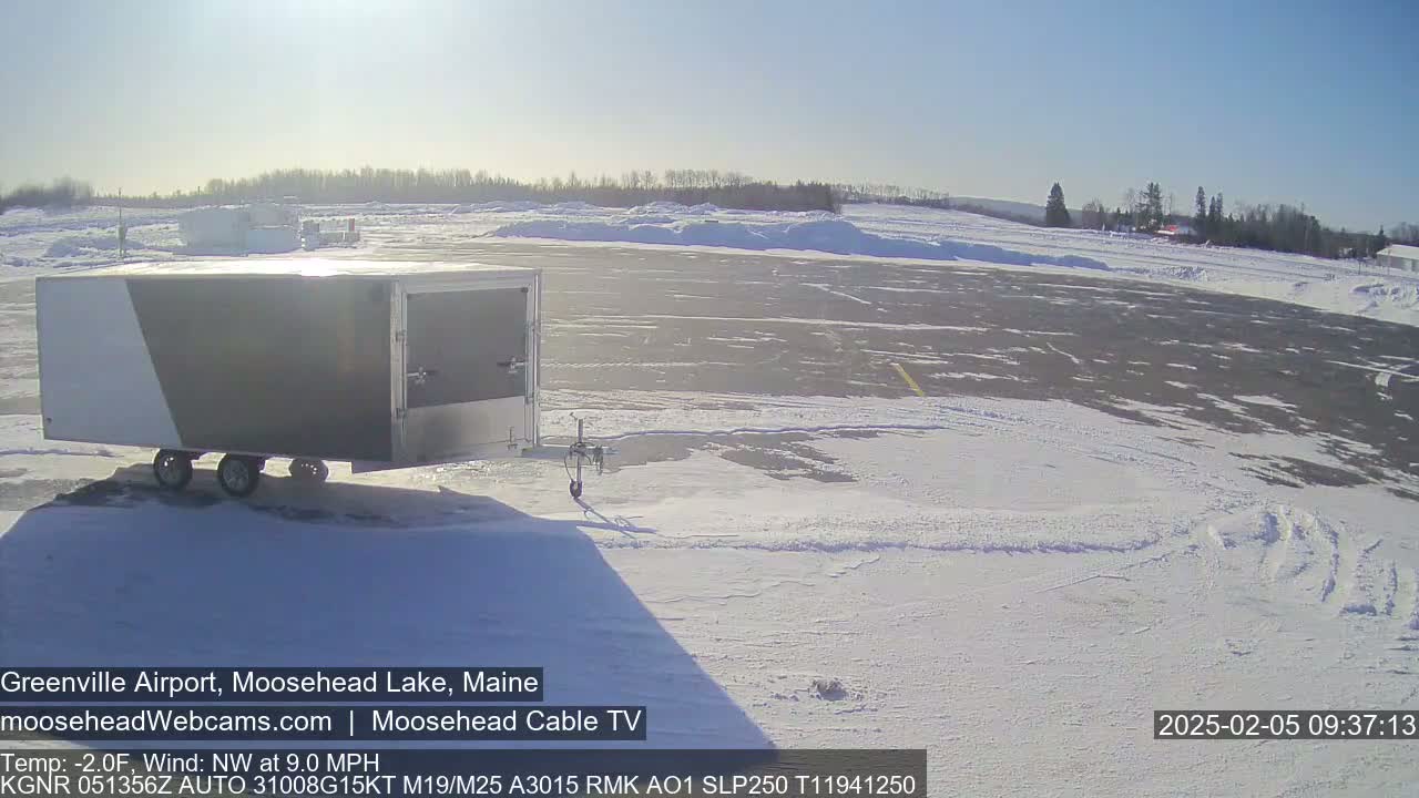 A gray and white enclosed trailer sits on a snow-covered tarmac under a sunny sky with a light northwest wind.