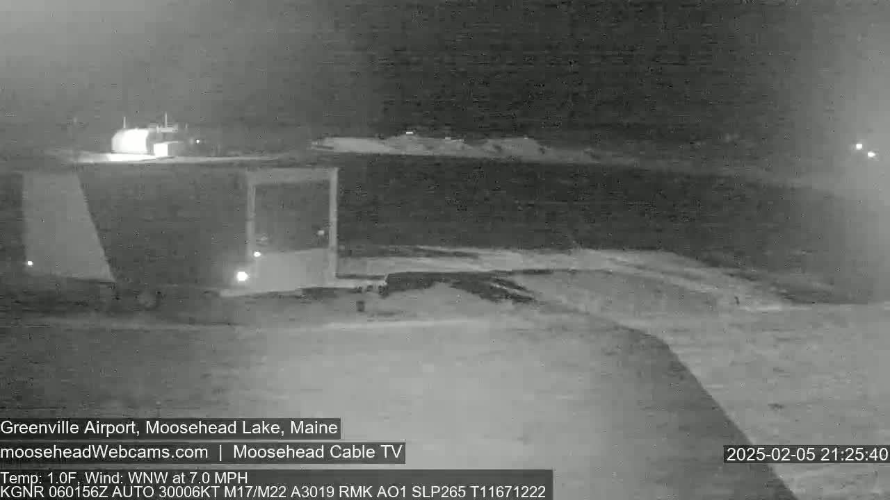 A nighttime, low-light image shows a dark, snow-covered landscape with a building and some faint lights in the distance.