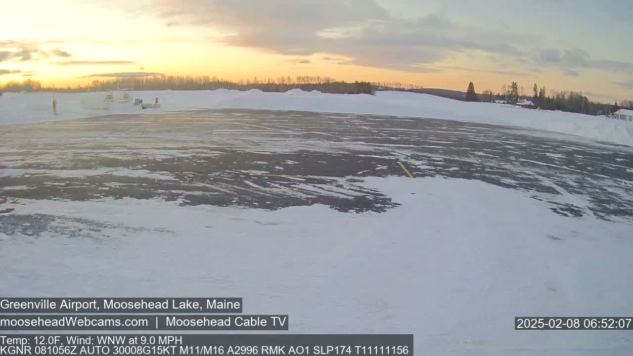 A snow-covered airport runway is visible at sunrise, with a few buildings and trees in the background.