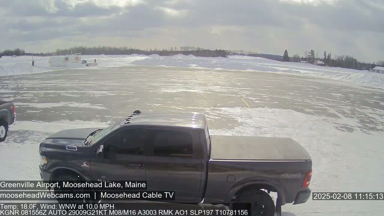 A dark-gray pickup truck is parked in a snowy airport lot under a partly cloudy sky.