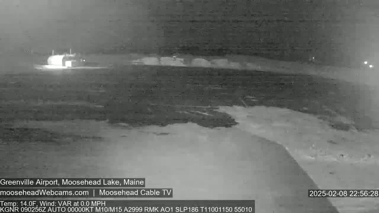 A nighttime, low-light grayscale image shows a snow-covered airport runway and a building, with calm weather conditions.