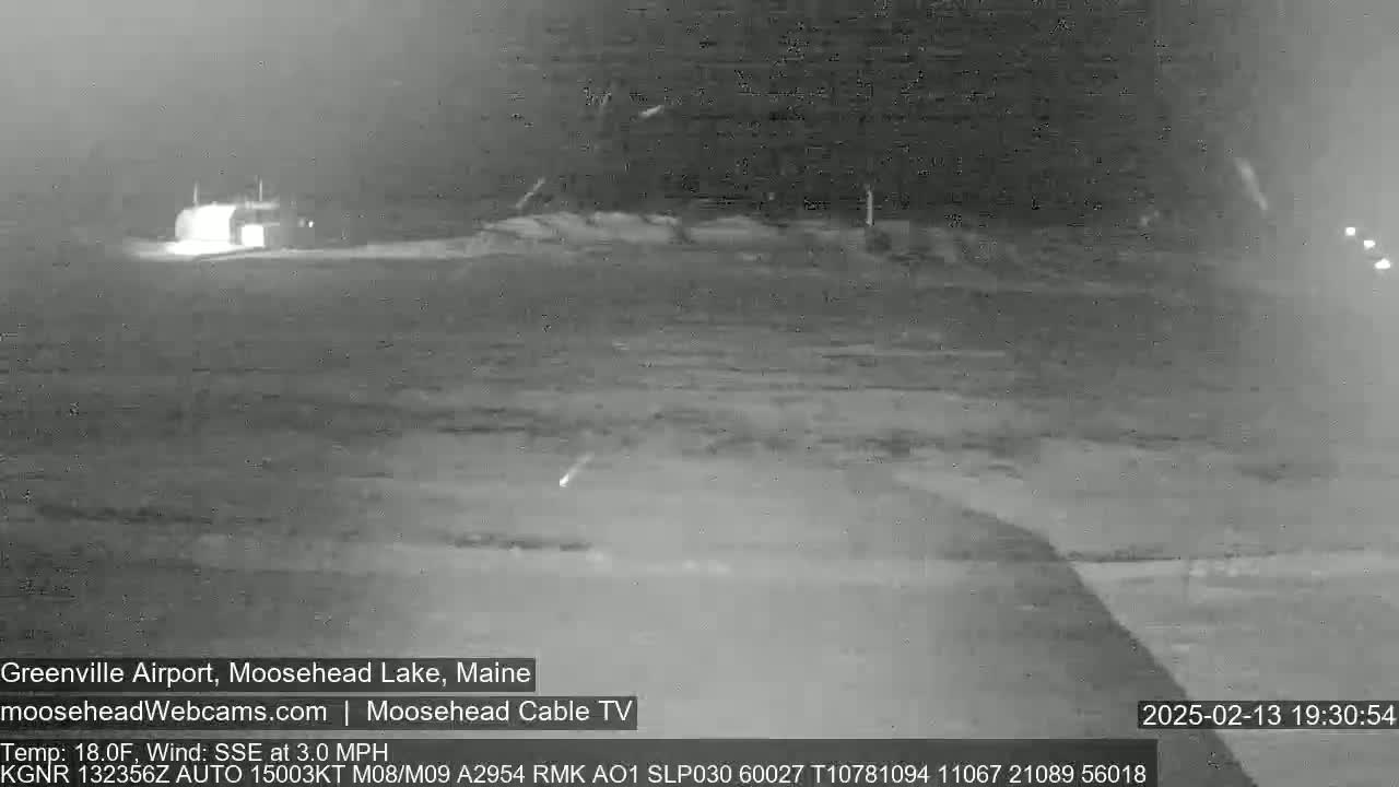 A nighttime, low-light grayscale image shows a mostly empty airfield with a building in the distance, under a dark sky with several light streaks possibly from meteors.
