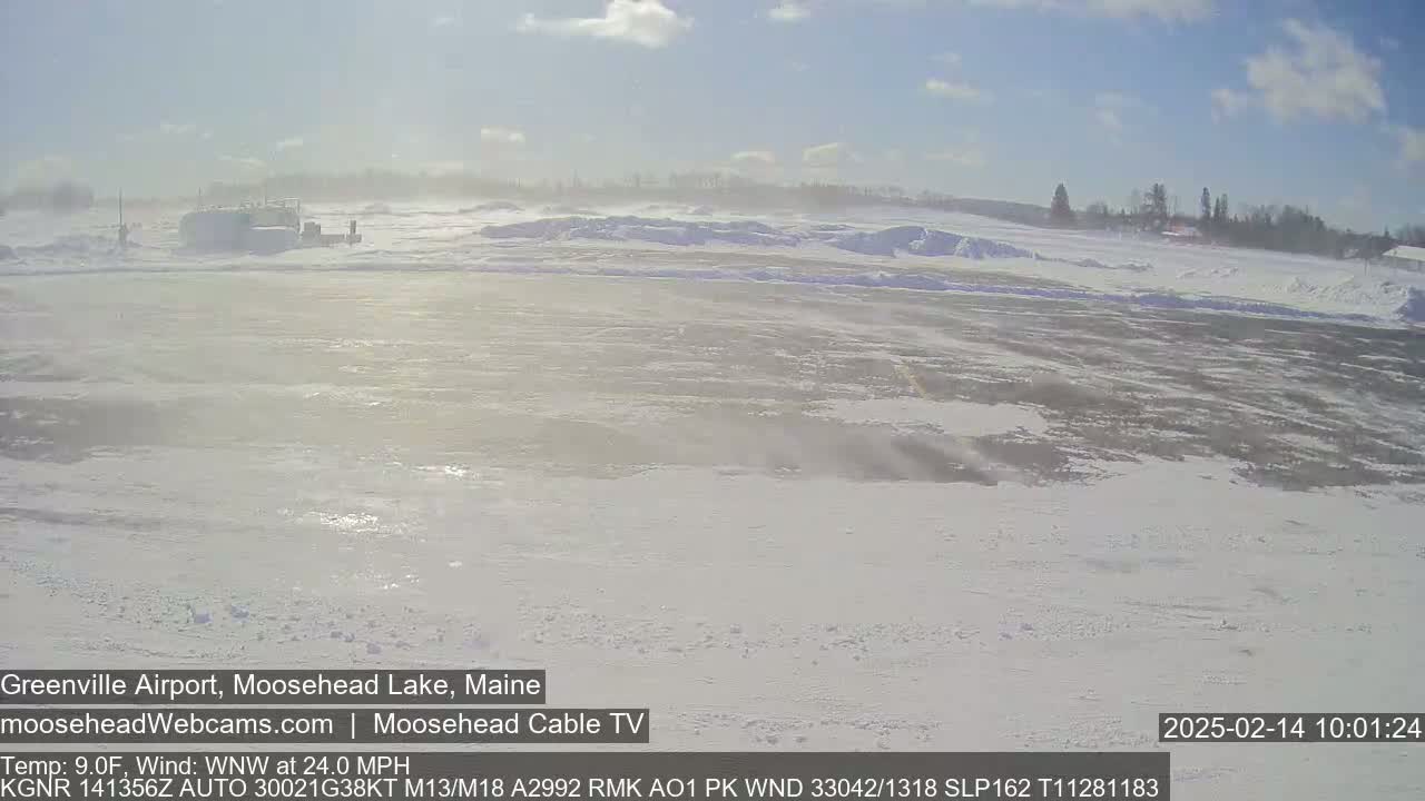 A snow-covered airfield is visible under a mostly sunny sky with a strong wind blowing snow across a flat, wide expanse.