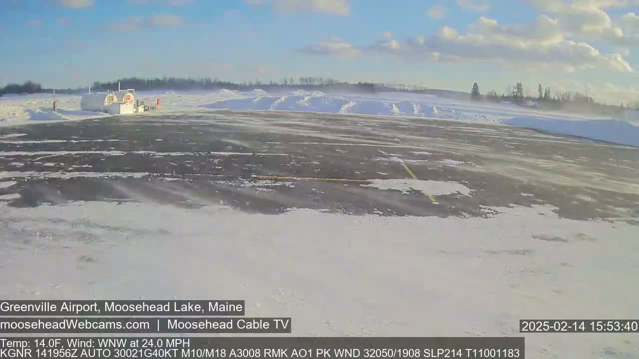 A snow-covered airport tarmac with a small building and snowdrifts in the background under a partly sunny sky with a strong wind.