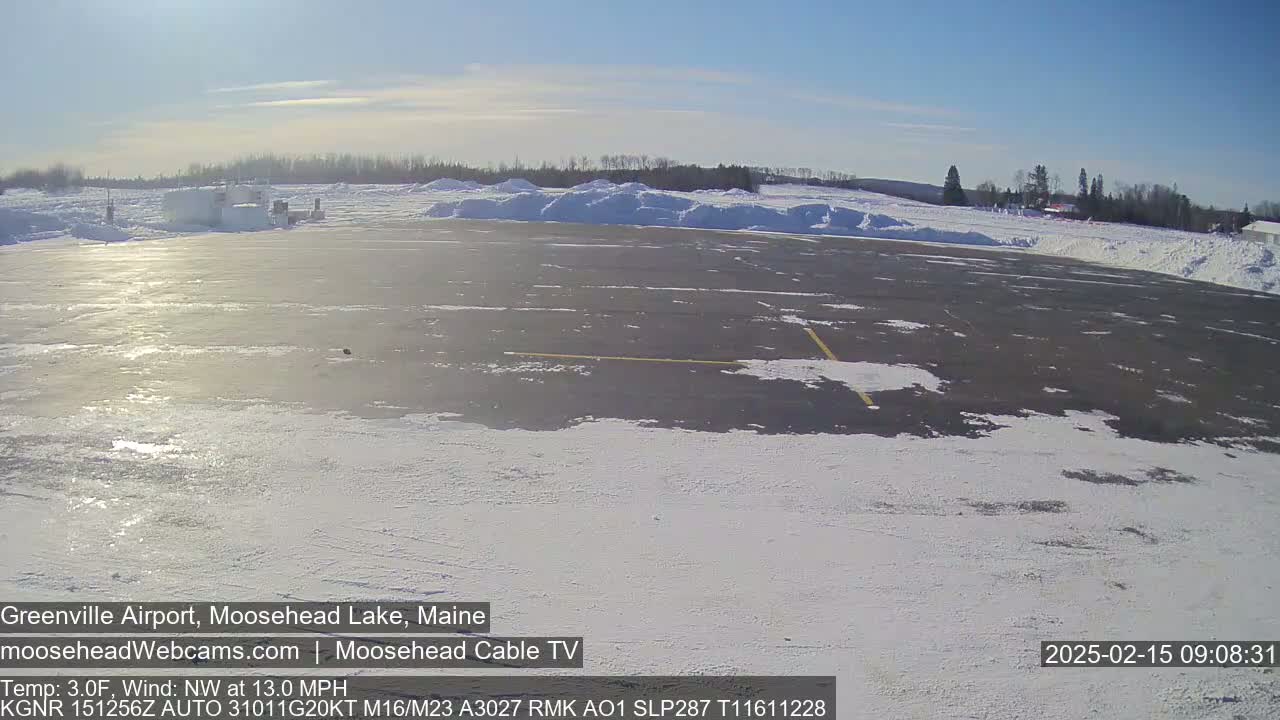A snow-covered airport tarmac is visible under a sunny sky, with snowdrifts along the sides and a treeline in the distance.