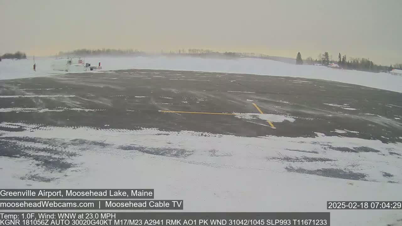 A snow-covered airport tarmac with a building in the distance, under a gray, overcast sky with strong winds.