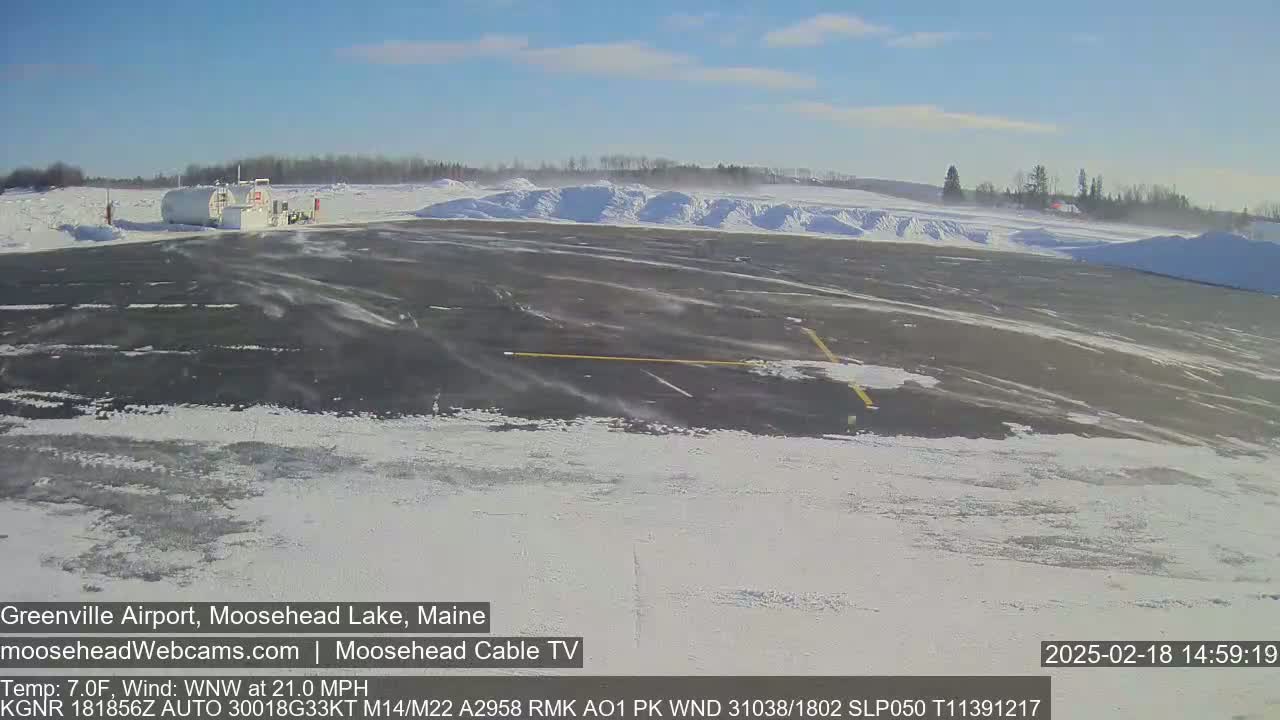 A snow-covered airport tarmac with a building in the background on a windy, sunny day.