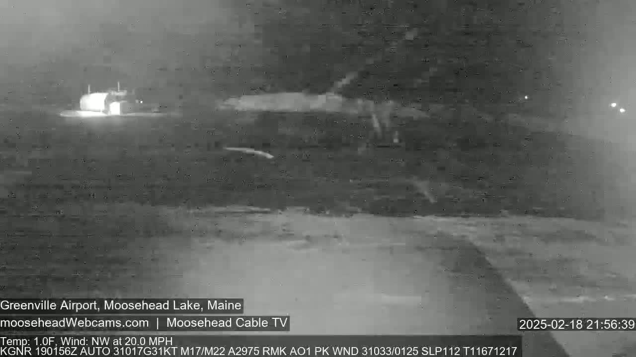 A nighttime, low-visibility scene shows a snowy or icy ground with a building faintly visible in the distance under a dark, possibly overcast sky, with a northwest wind blowing at 20 mph.