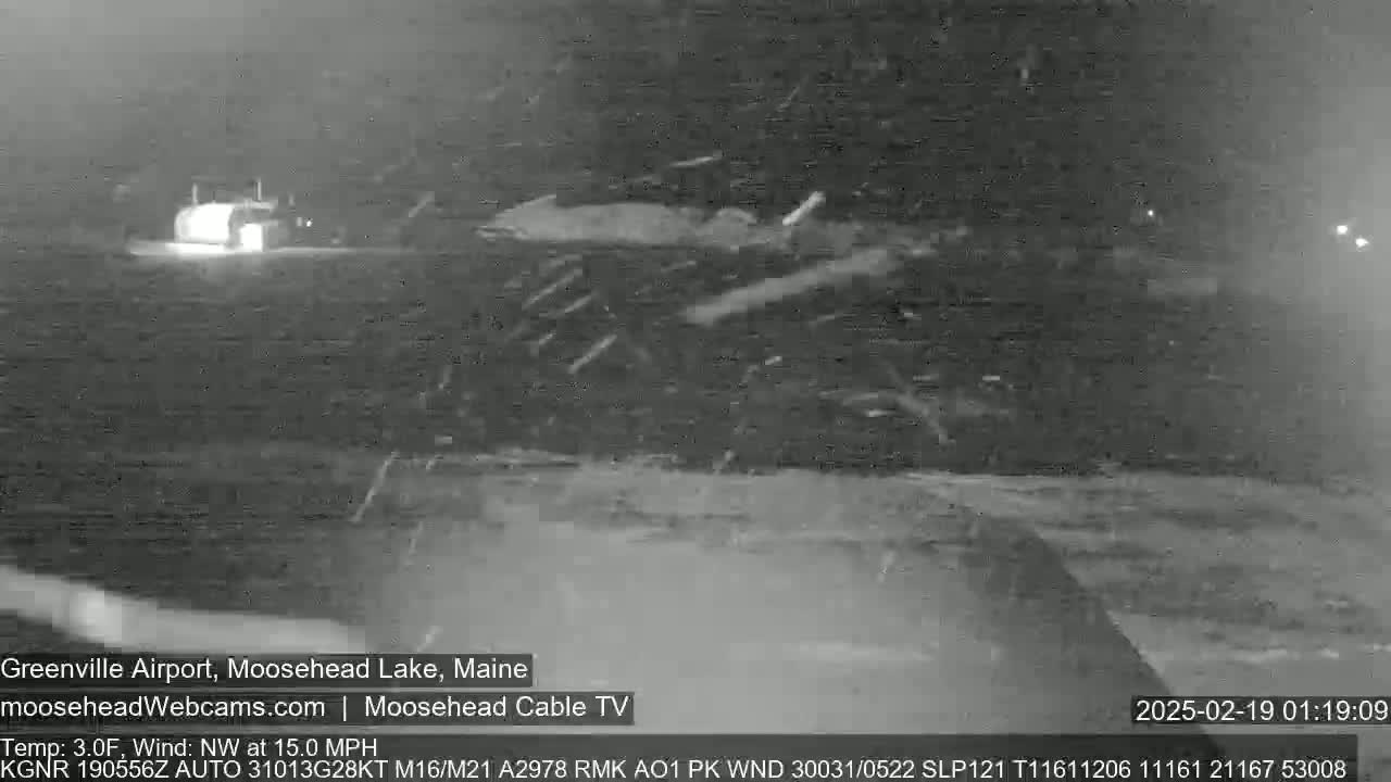 A nighttime, snowy scene shows a building in the distance with snow blowing across a flat, open area.