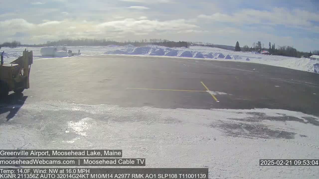 A partly sunny day shows a snow-covered airport tarmac with a snow plow and piles of snow in the background.