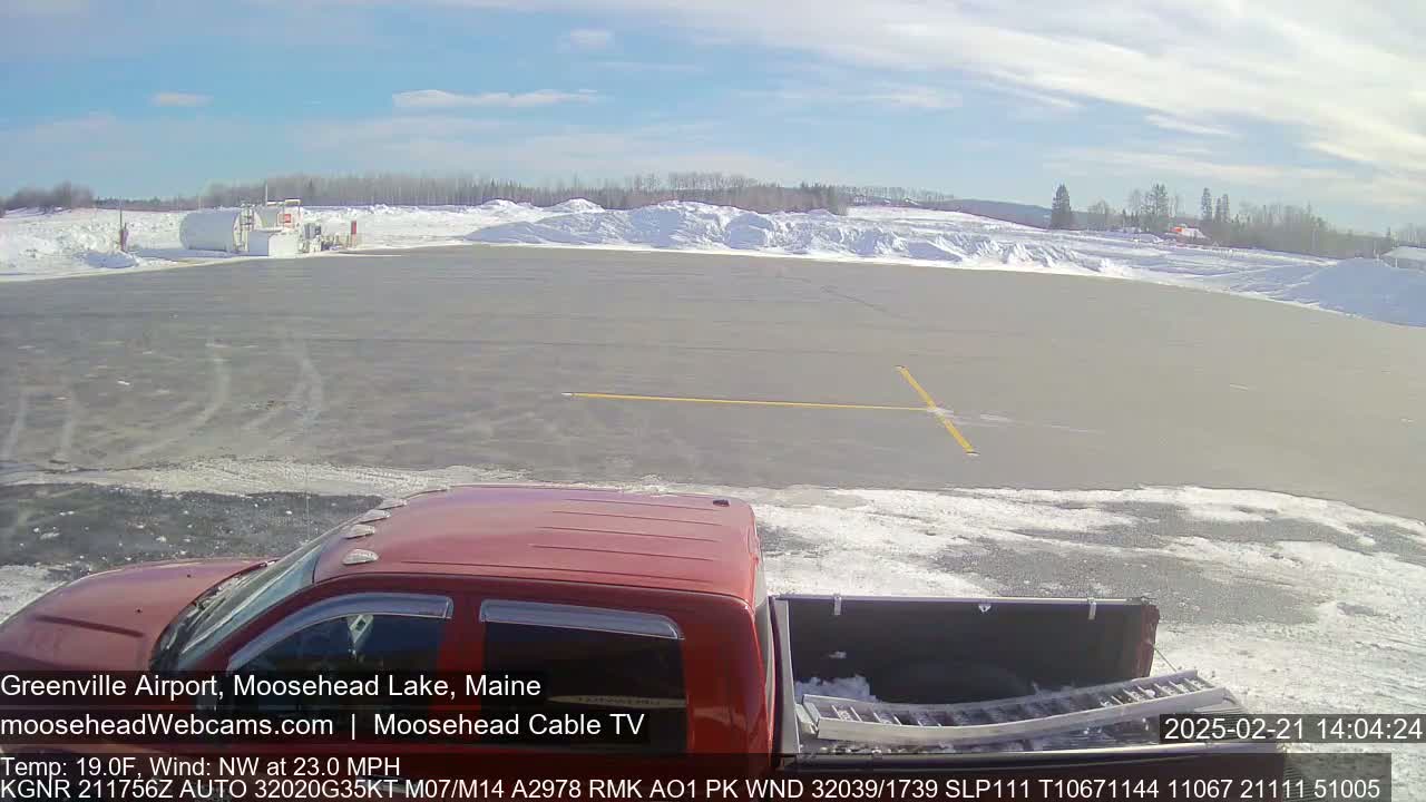 A red pickup truck with a black truck bed is parked on a snow-covered tarmac on a sunny day with a light wind.