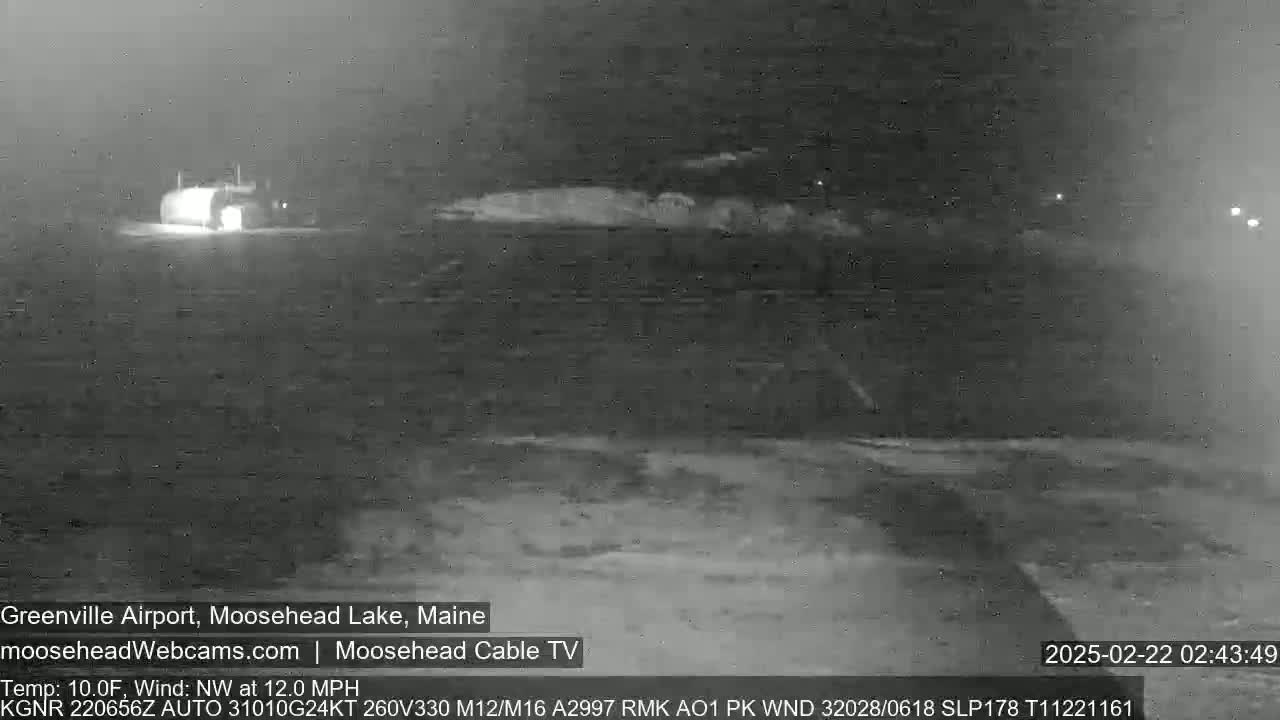 A nighttime, low-light view of a snow-covered airfield with a building in the distance, under a dark sky with a northwest wind blowing at 12 mph and a temperature of 10 degrees Fahrenheit.