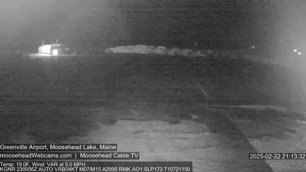 A nighttime, low-visibility view of a snow-covered landscape showing a building and a road under a dark sky.