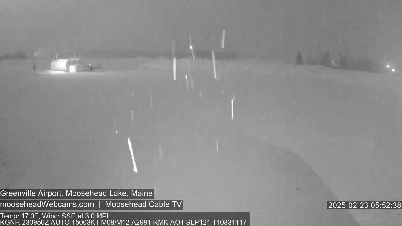 A nighttime, snowy scene shows a building and a flat expanse of land under a lightly falling snow.
