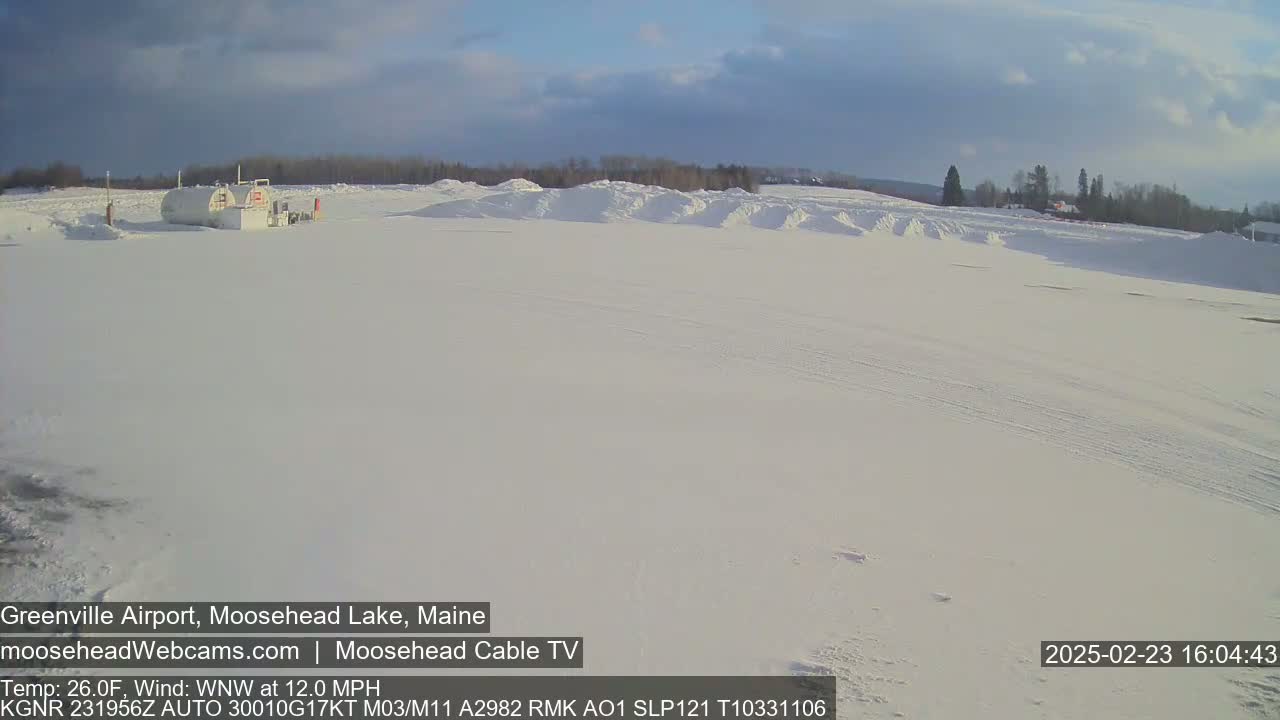 A snow-covered airfield with a few fuel tanks and distant trees under a partly cloudy sky.