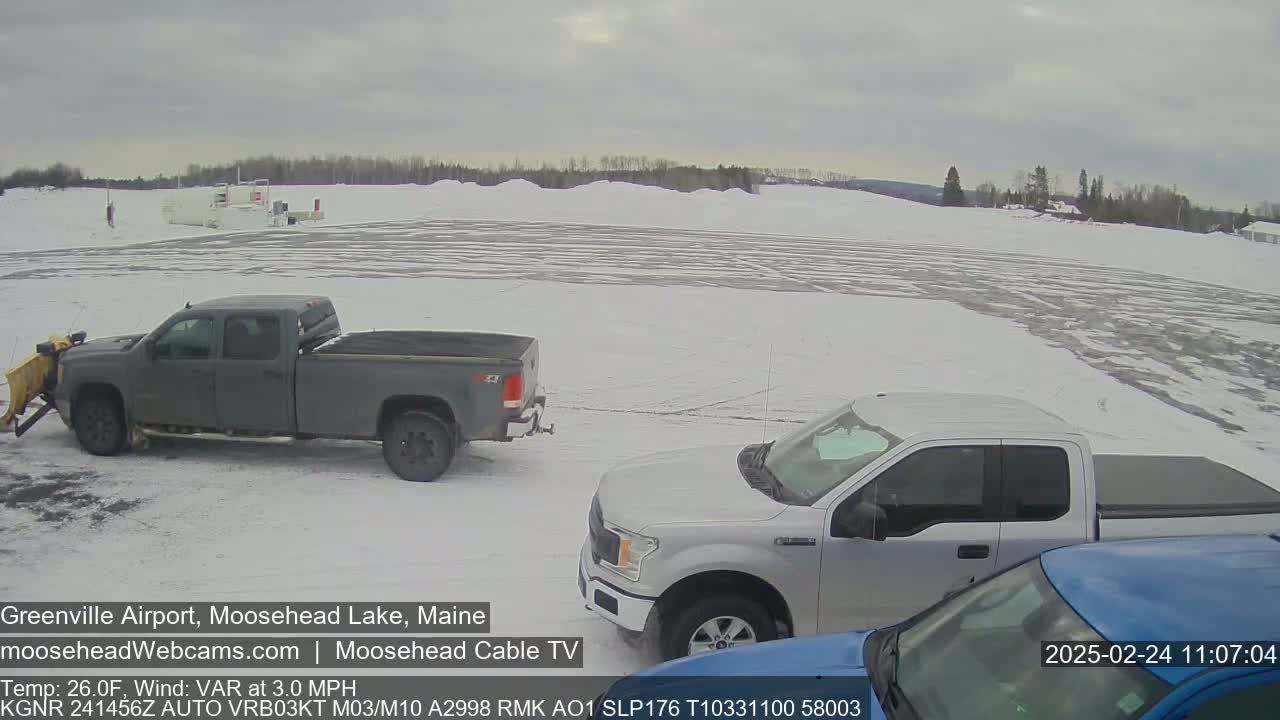 A snow-covered airport with two pickup trucks parked and a third partially visible, under an overcast sky.