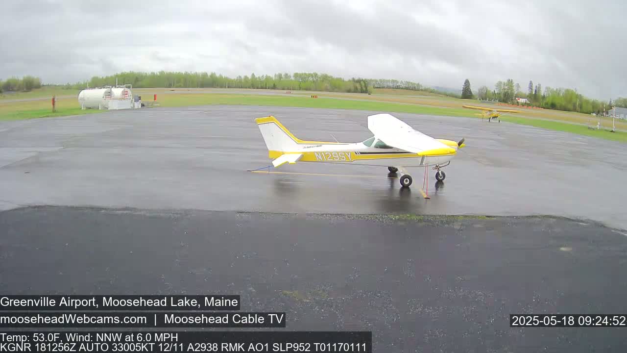 A wet airport tarmac shows two small airplanes parked, one in the foreground and another further back, under an overcast sky.