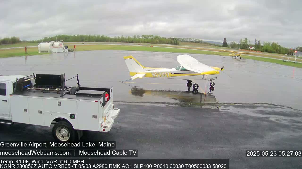 A small yellow and white airplane is parked on a wet airport tarmac next to a white pickup truck, with another smaller airplane visible in the background under overcast skies.