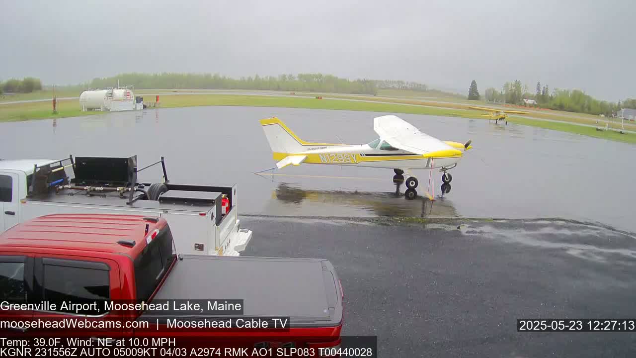 A yellow and white airplane is parked on a wet tarmac next to two pickup trucks in rainy weather.