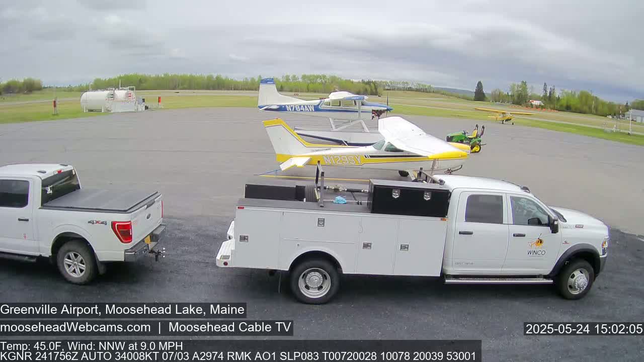 A white pickup truck and service van are parked at an airport on an overcast day next to two small airplanes and a third farther away; a fuel tank is visible in the background.