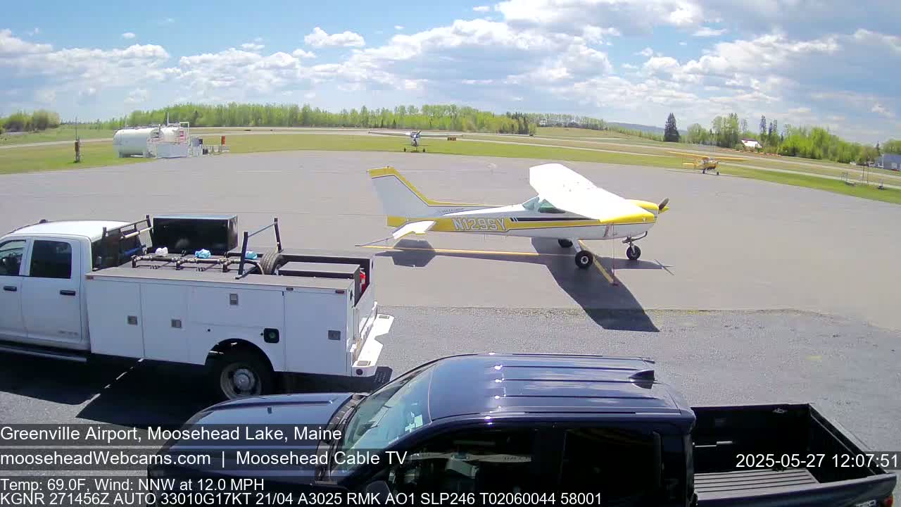 A yellow and white single-engine airplane is parked on an airport tarmac under a partly cloudy sky, near a white pickup truck and a dark-colored SUV.