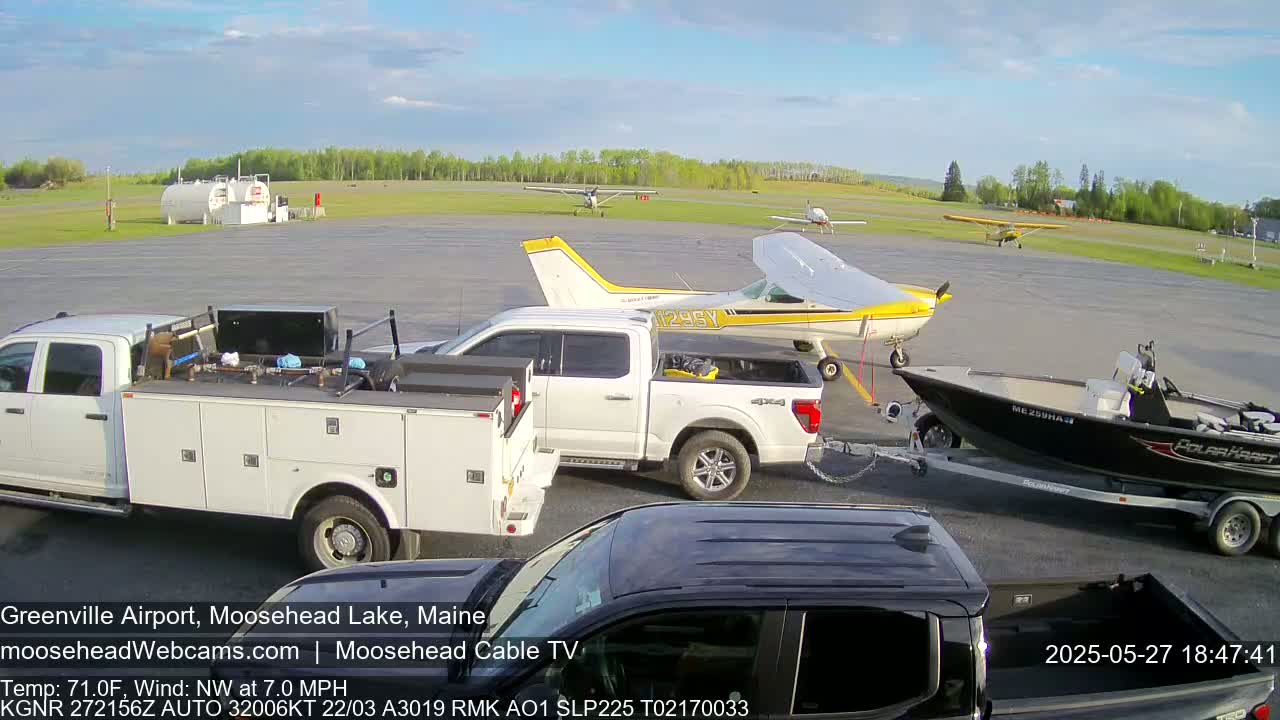 A sunny airport scene shows several small airplanes, a white pickup truck towing a boat on a trailer, and a white service truck.