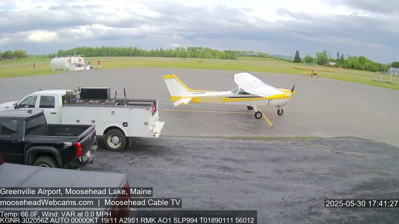 A yellow and white single-engine airplane is being towed by a white pickup truck on an airfield under a partly cloudy sky.
