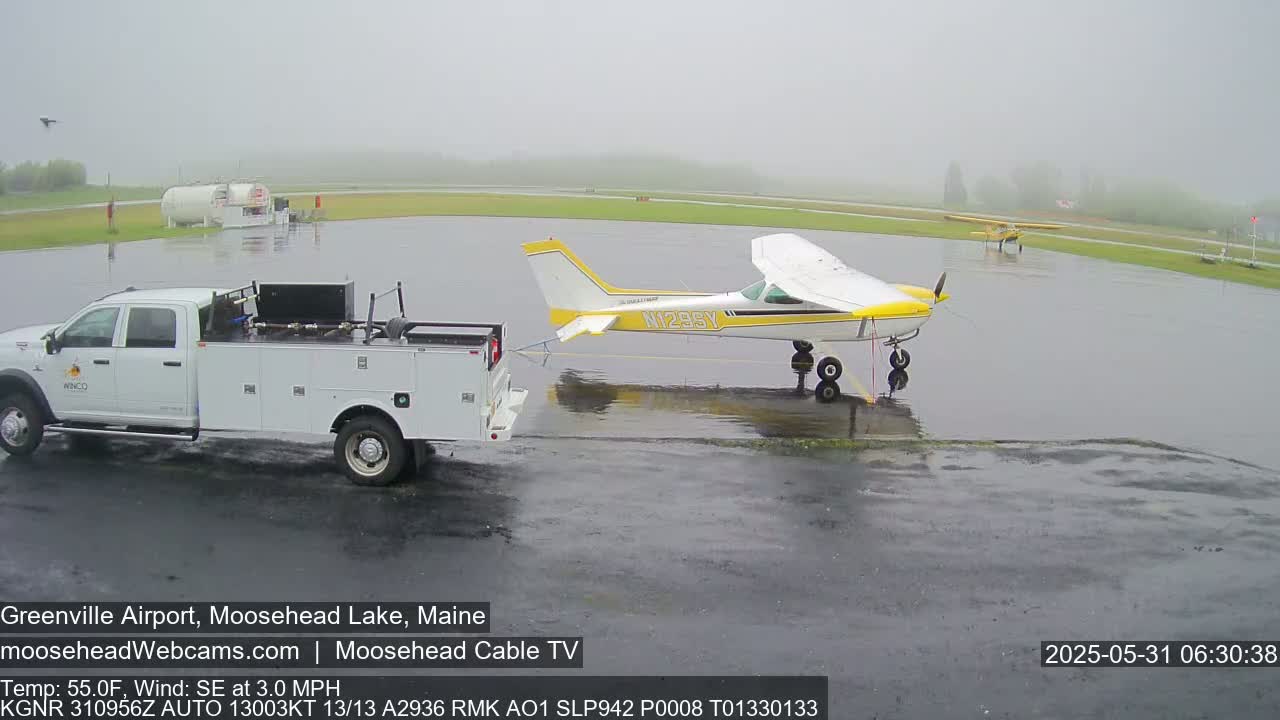 A white pickup truck tows a yellow and white airplane on a wet airport tarmac in foggy conditions.