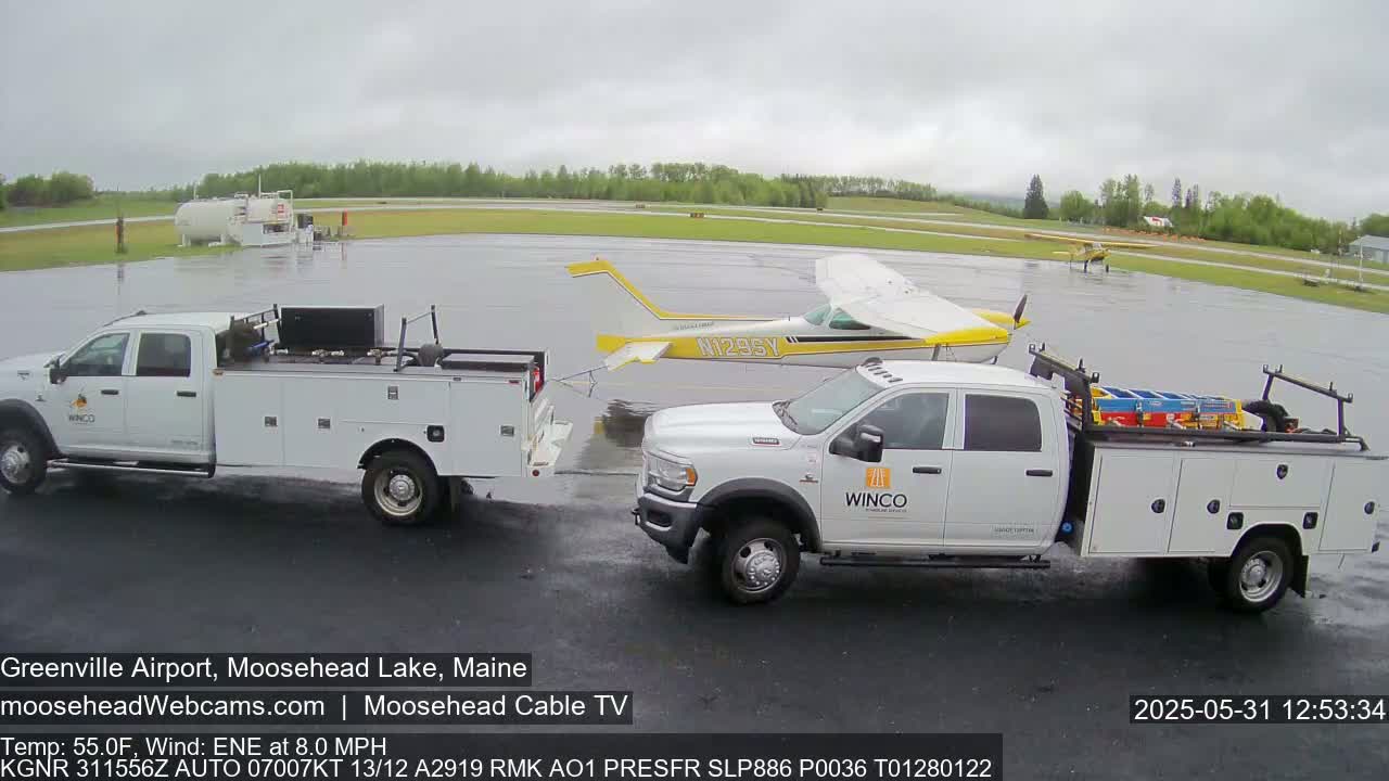 A small yellow airplane is parked on a wet airport tarmac next to two white pickup trucks on a cloudy day.