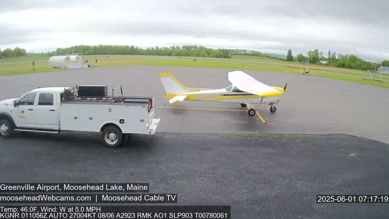 A white pickup truck is towing a yellow and white airplane on an airfield under an overcast sky.