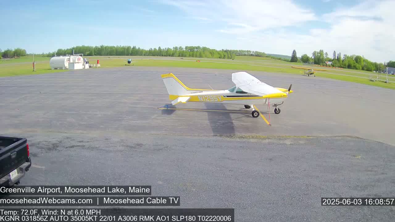 A small airplane sits on a tarmac at an airport on a partly sunny day, with another plane and a fuel truck visible in the background.