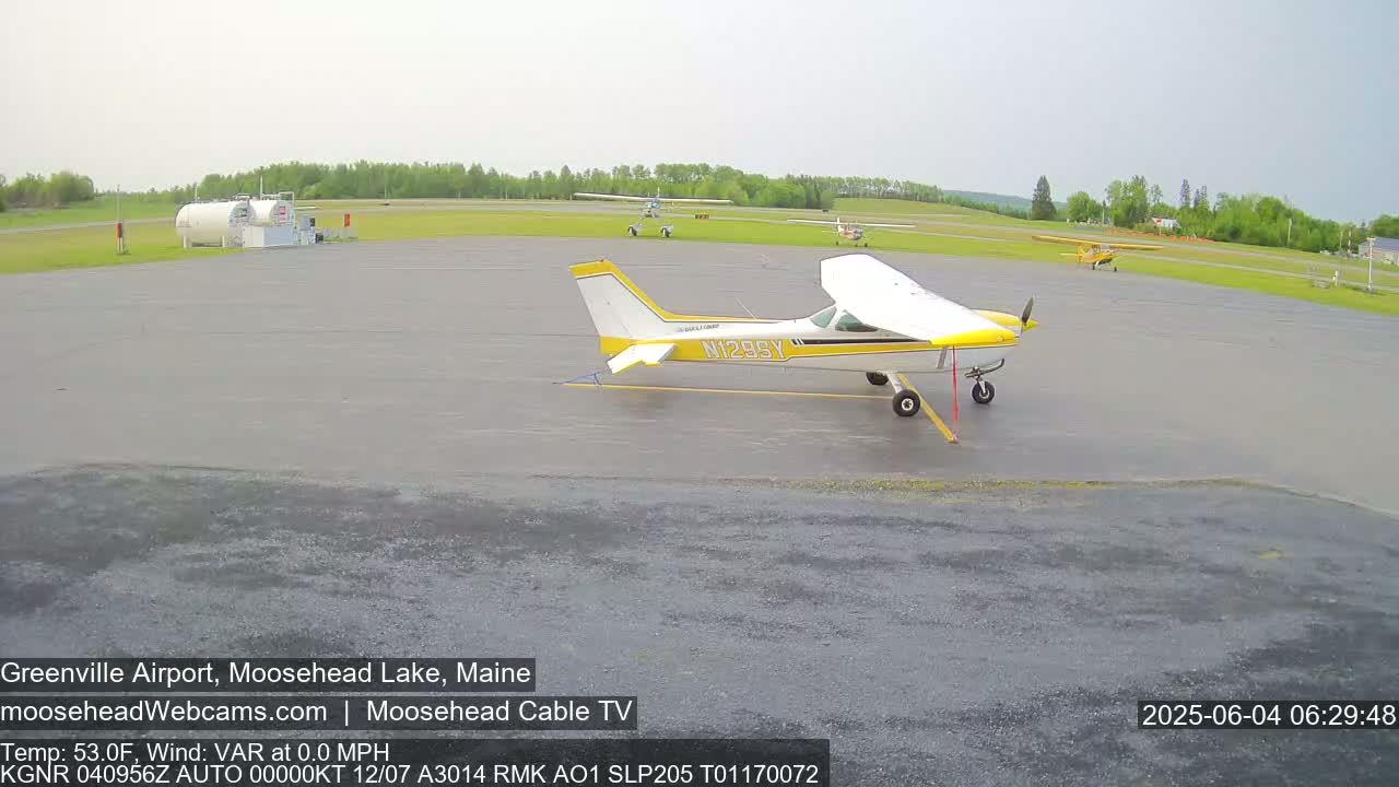 A yellow and white small airplane sits on a wet tarmac at an airport surrounded by other smaller planes and a fuel station, under an overcast sky.