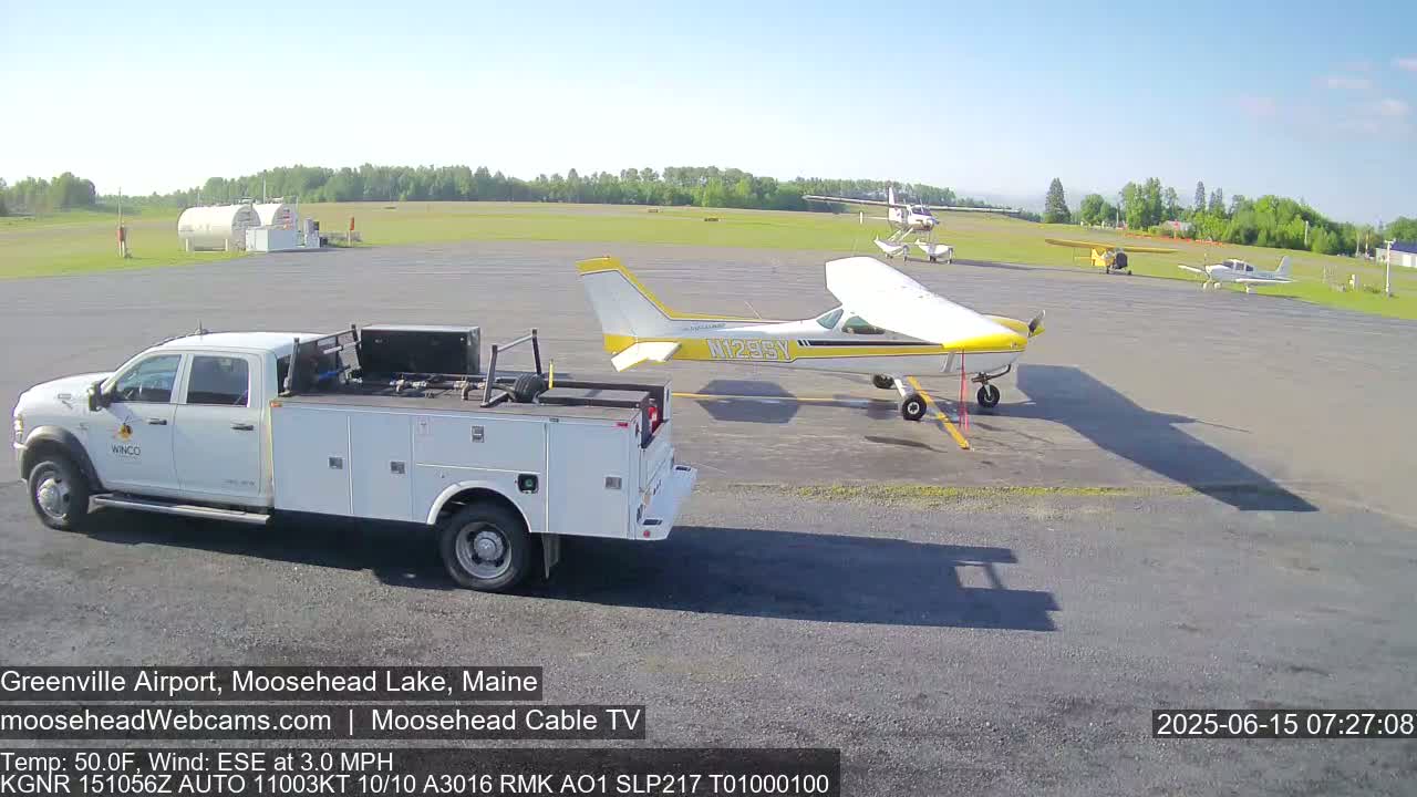 A white pickup truck is parked on an airport tarmac next to a yellow and white single-engine airplane; two other small planes are parked further away under a clear sky.