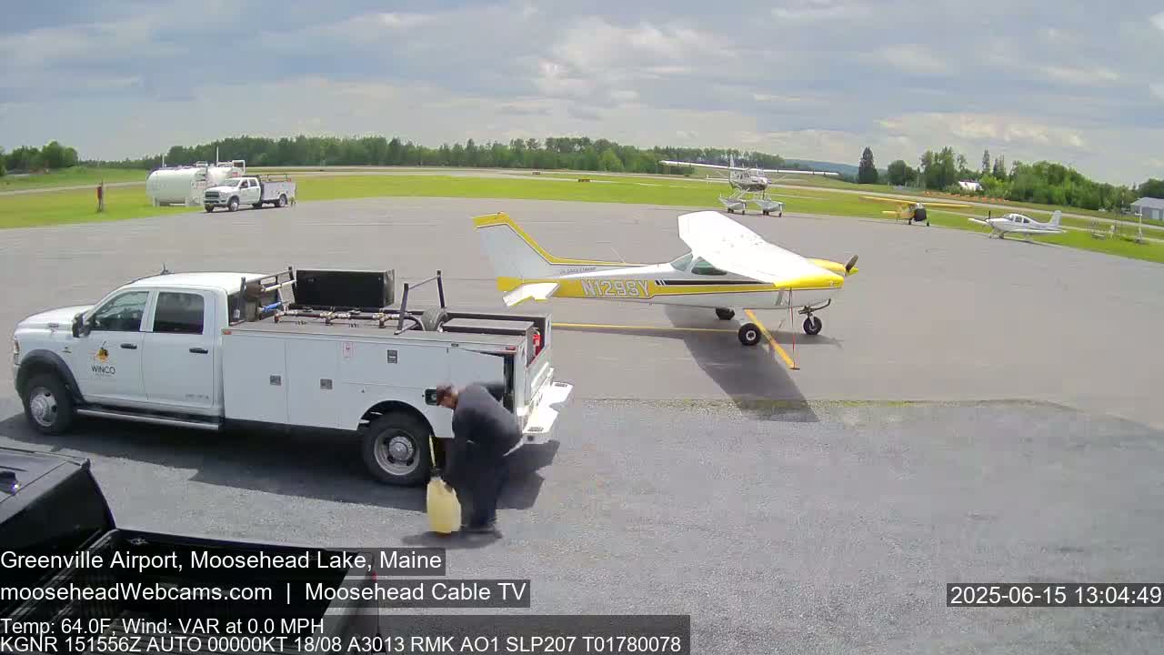 A person is refueling a small yellow and white airplane on a tarmac, with other planes and trucks parked nearby under partly cloudy skies.