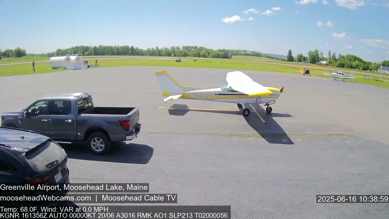 A small airport tarmac on a sunny day with a yellow and white single-engine airplane, two other smaller planes parked in the distance, a gray pickup truck, and a portion of a dark-colored SUV.
