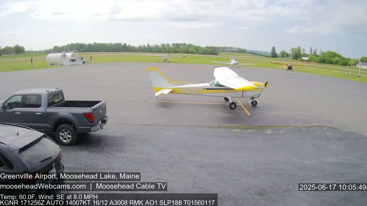 A small airport tarmac under partly cloudy skies shows several small airplanes parked, a pickup truck, and another vehicle partially visible.