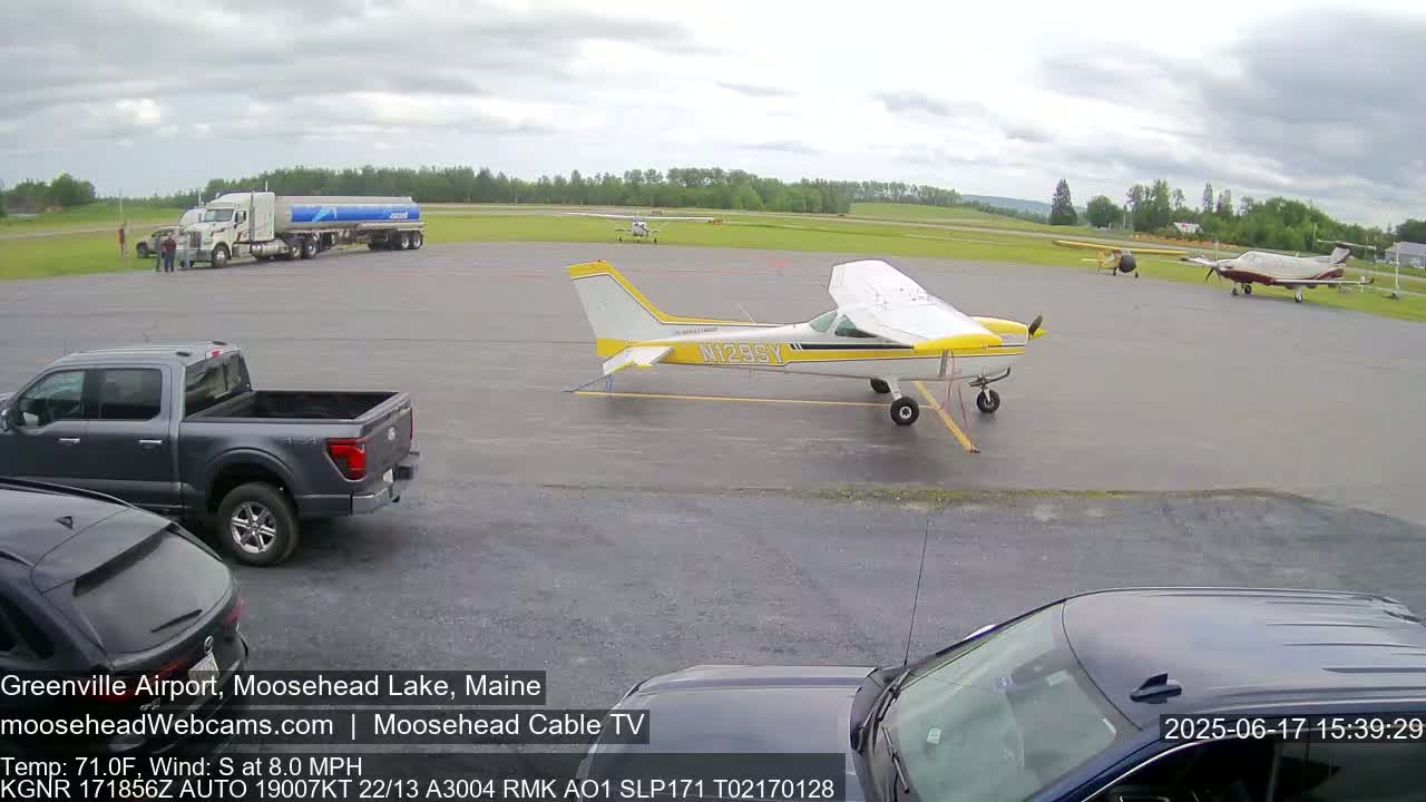 An airport tarmac on an overcast day shows several small airplanes, a fuel tanker, a pickup truck, and several cars.