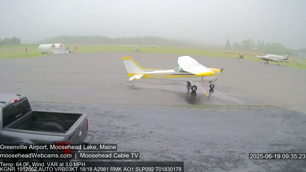 A wet airport tarmac with several small airplanes parked, a pickup truck, and a large propane tank visible under an overcast sky.