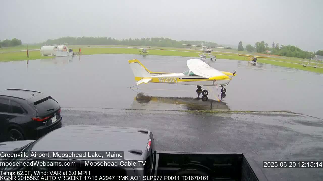 A rain-soaked airport tarmac with several small airplanes parked, one prominently featured in the foreground, and cars parked nearby.