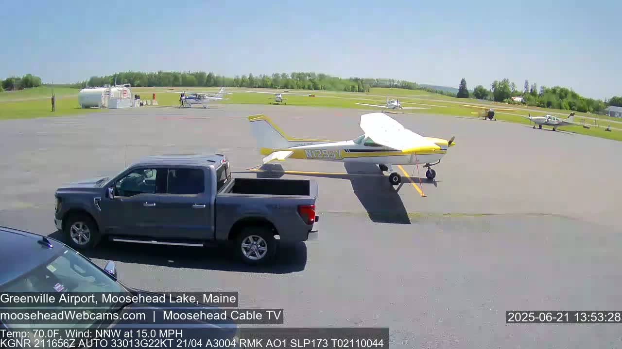 A gray pickup truck is parked next to a yellow and white single-engine airplane on a sunny airport tarmac with several other planes visible.