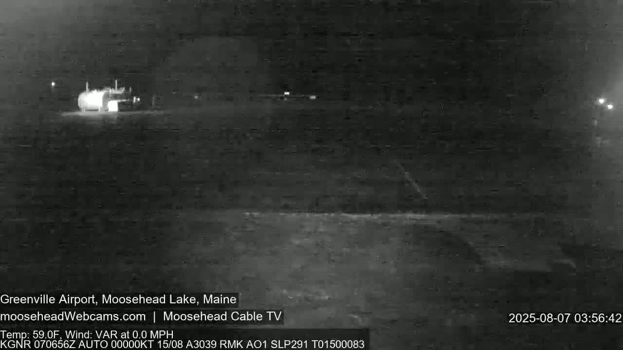 A nighttime, low-light infrared image shows a sparsely lit airfield with a single vehicle near a building.
