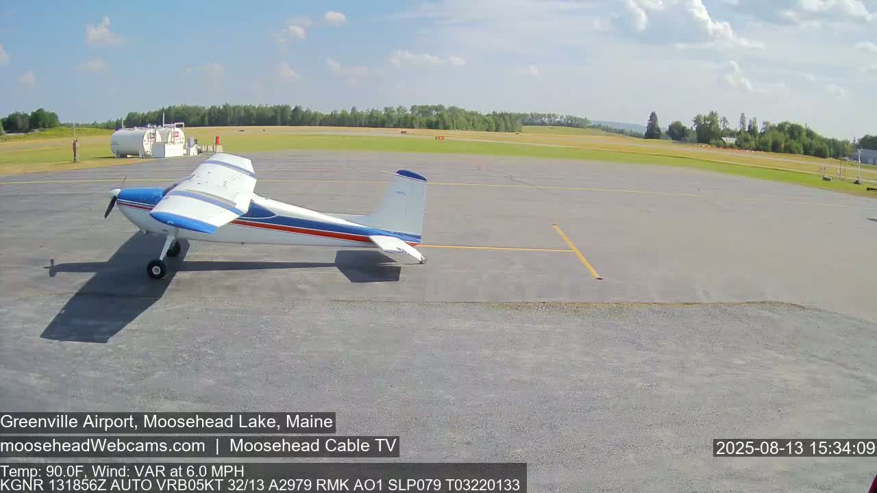 A small, single-engine airplane sits on a tarmac at an airport on a partly cloudy day with a temperature of 90 degrees Fahrenheit and a variable wind of 6 mph.