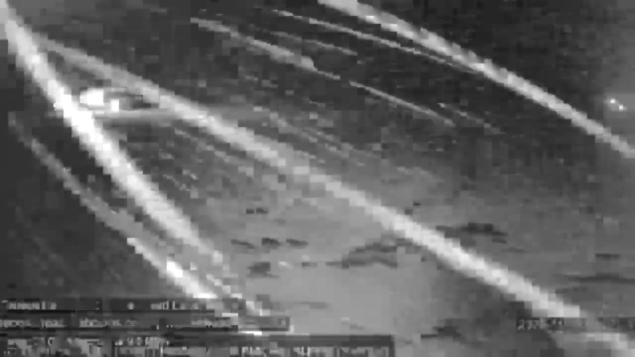 A grainy, black-and-white image displays an outdoor night scene featuring numerous bright, diagonal streaks of light, likely from moving vehicles on a road, under clear conditions.