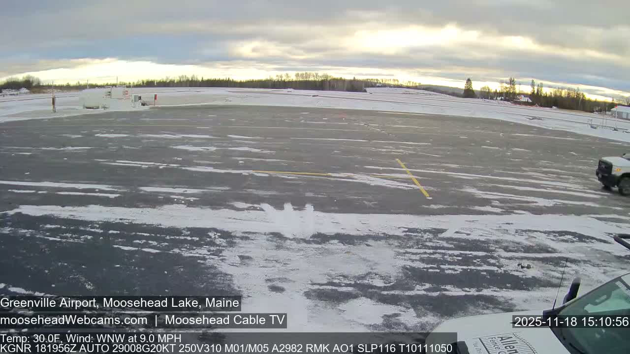 From the perspective of a foreground vehicle, a snow-dusted airport tarmac with yellow markings extends under a partly cloudy sky, featuring distant buildings, a line of trees on the horizon, and another vehicle driving on the right, on a cold, wintery day.