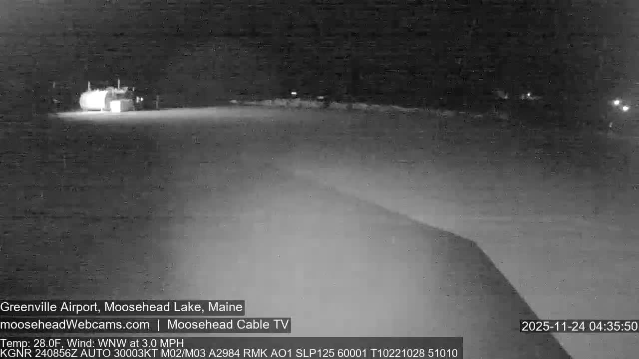 A grayscale night scene captures a wide, flat expanse, possibly a road or airfield, with a brightly illuminated building or vehicle visible in the distant left and smaller lights scattered along the horizon, all under a clear sky.