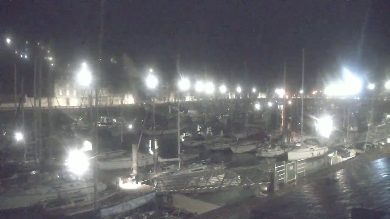 Many sailboats are docked at a marina at night under bright lights.