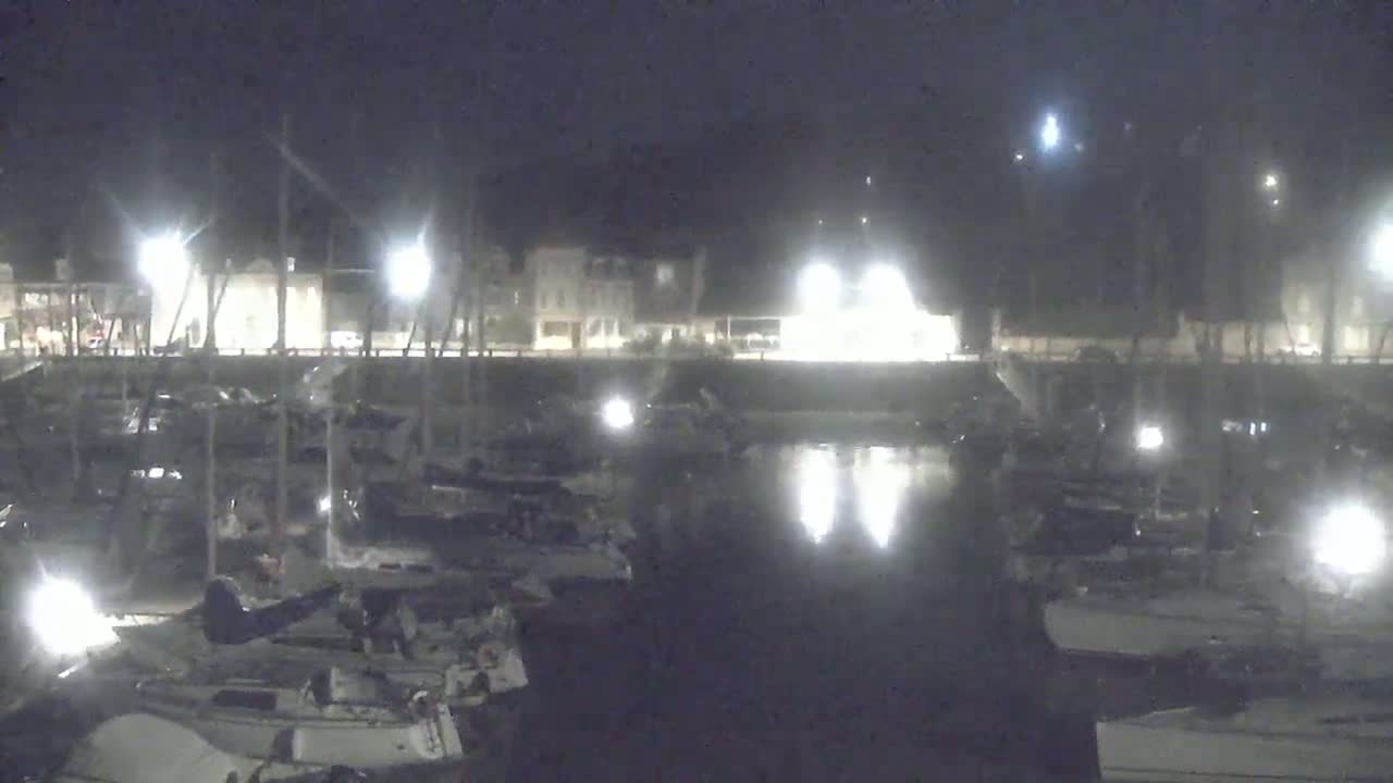 At night, many boats are docked in a marina, with lights illuminating the water and the buildings along the shore.