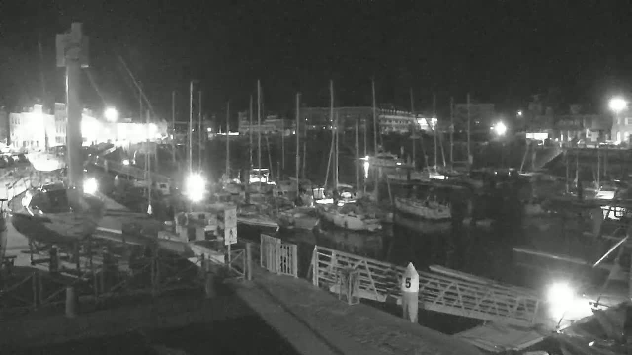 A nighttime, black-and-white view of a marina filled with docked sailboats and illuminated buildings along the waterfront.