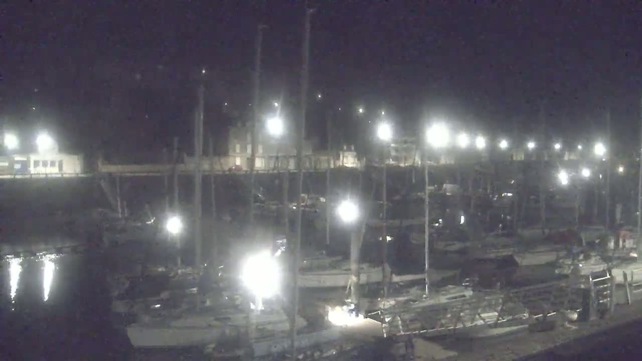 Many sailboats are docked at a marina at night under bright artificial lights.