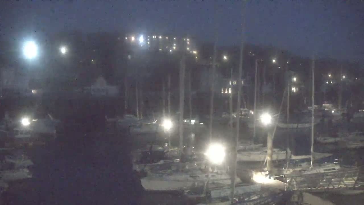 A nighttime view of a marina with numerous sailboats moored, illuminated by scattered lights under a dark, possibly hazy sky.